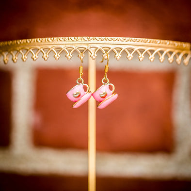 small pink tea cup and saucers earrings with hearts dangling from display on brick background