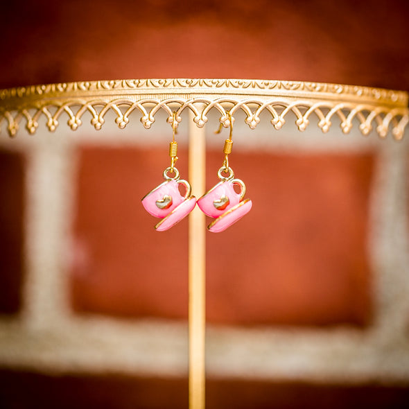 small pink tea cup and saucers earrings with hearts dangling from display on brick background