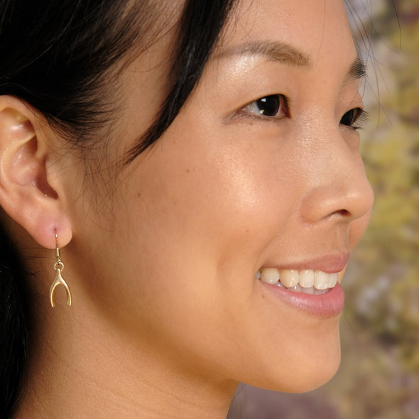 Beautiful model wearing gold dangle wishbone earrings smiling in front of tree and flower backdrop